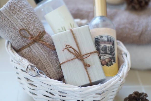 White wicker basket with spa items including folded towel, soap, pump bottle, and perfume spray, surrounded by pinecones on tile surface