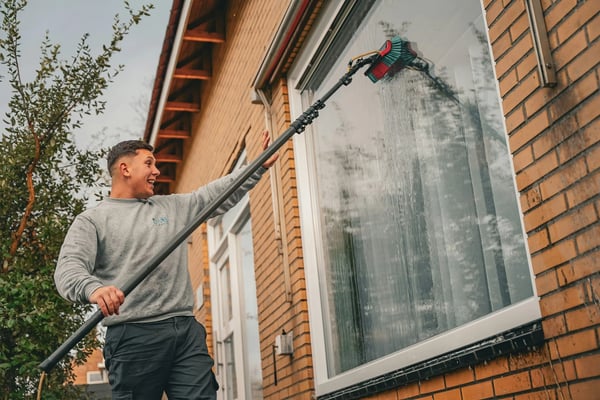 Man using long-handled brush to clean exterior brick wall and window of residential building
