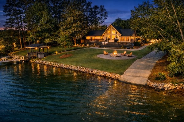 Waterfront house at dusk with illuminated windows, fire pit, Adirondack chairs, dock, and wooded grounds along calm water.